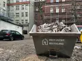Kontener na odpady budowlane, kod odpadu 17 09 04. Wypełniony gruzem i opakowaniami, obok stoi czarny samochód.