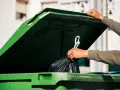 Ręka wrzuca worek ze śmieciami do zielonego kontenera. Zastanawiasz się, ile kosztuje wywóz śmieci?