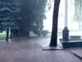 Intensywny deszcz nawalny zalewa parkową alejkę i trawnik. Drzewa pochylają się pod naporem wody.