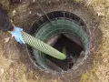 Ręka w niebieskiej rękawiczce wprowadza wąż do szamba betonowego. Drenaż szamba jest kluczowy dla prawidłowego funkcjonowania.