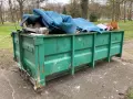 Zielony kontener KP7 wypełniony po brzegi niebieską wykładziną i innymi odpadami.