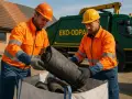 Dwóch pracowników w pomarańczowych kurtkach i kaskach wyjmuje z worka zrolowane materiały. Obok stoi zielona śmieciarka z napisem "EKO-ODPADY". To pszok papa, który trafi do recyklingu.