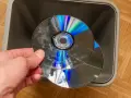 Dłoń trzyma pękniętą płytę CD/DVD nad szarym koszem na śmieci.