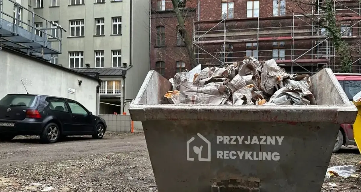 Kontener na odpady budowlane, kod odpadu 17 09 04. Wypełniony gruzem i opakowaniami, obok stoi czarny samochód.
