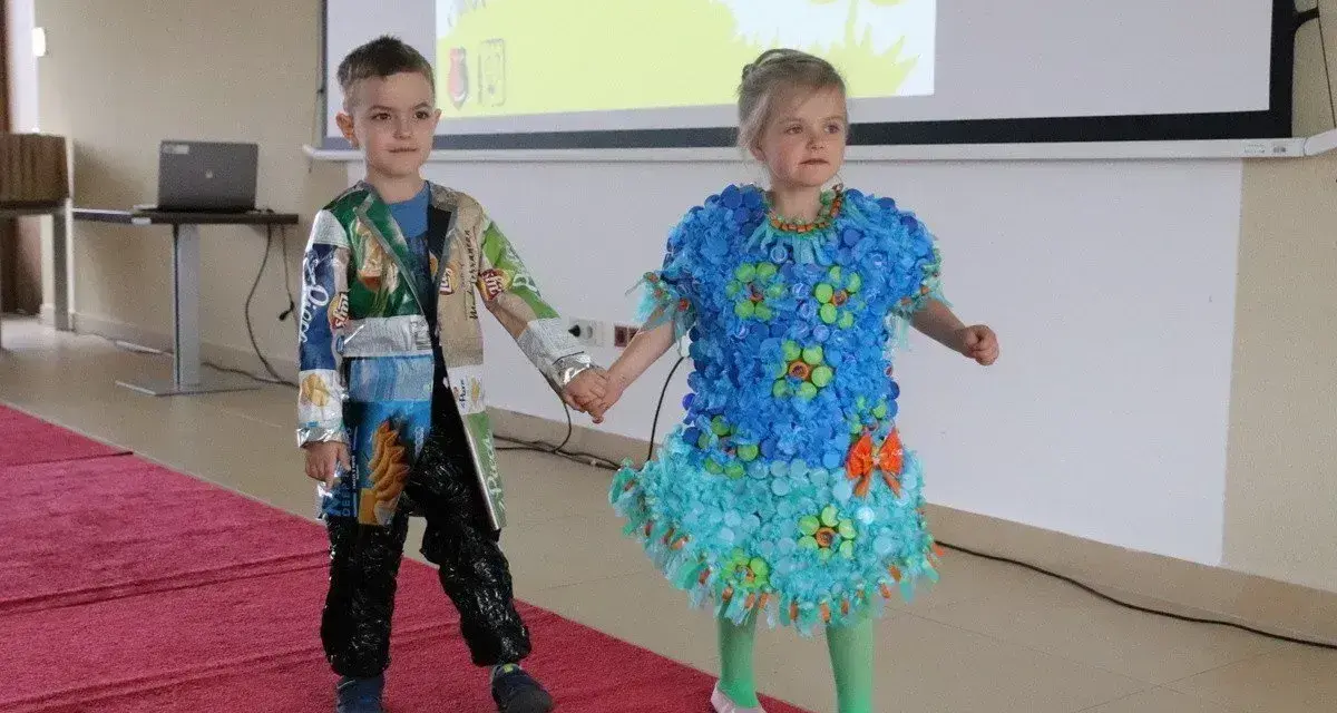 Chłopiec prezentuje strój z recyklingu, wykonany z opakowań po przekąskach. Jego koleżanka ma sukienkę z nakrętek.