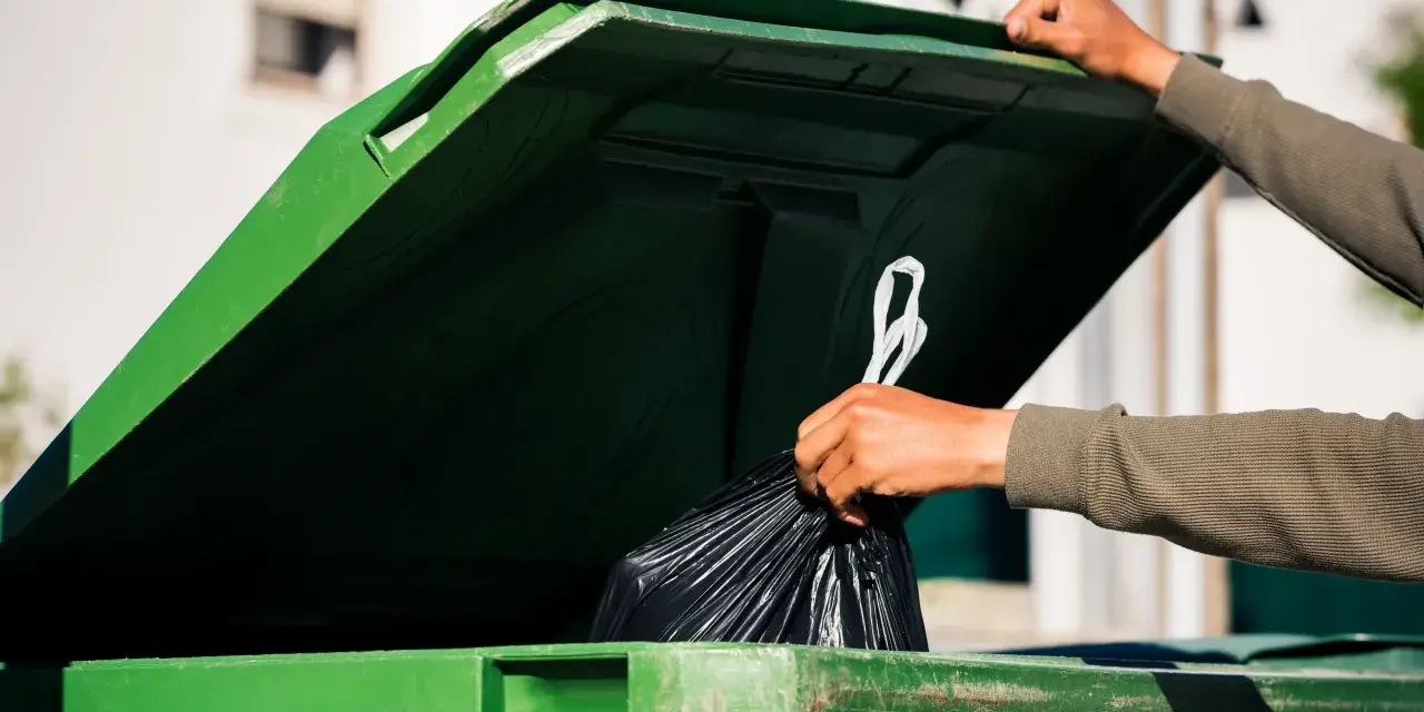 Ręka wrzuca worek ze śmieciami do zielonego kontenera. Zastanawiasz się, ile kosztuje wywóz śmieci?