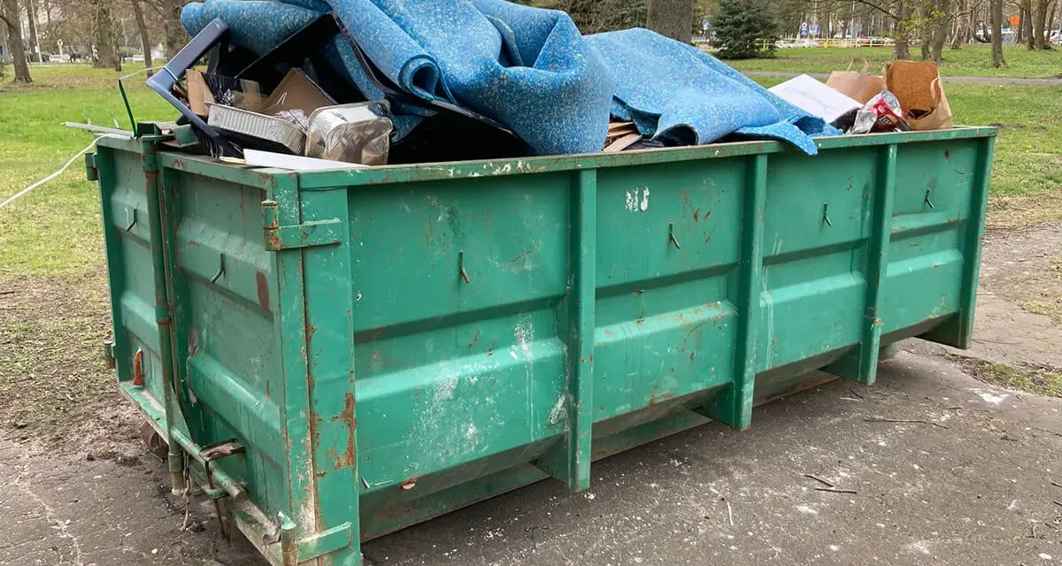 Zielony kontener KP7 wypełniony po brzegi niebieską wykładziną i innymi odpadami.