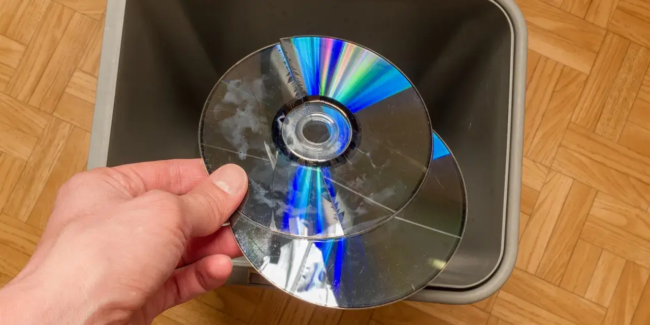Dłoń trzyma pękniętą płytę CD/DVD nad szarym koszem na śmieci.