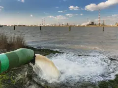 Rurociąg zrzuca wodę do rzeki, tworząc pianę. W tle widać przemysłowy krajobraz z rafinerią, gdzie mogą być używane surfaktanty niejonowe.