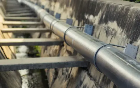 Metalowa rura biegnie wzdłuż betonowego koryta. To element kanalizacji ciśnieniowej, transportującej wodę.