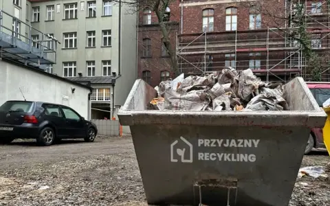 Kontener na odpady budowlane, kod odpadu 17 09 04. Wypełniony gruzem i opakowaniami, obok stoi czarny samochód.