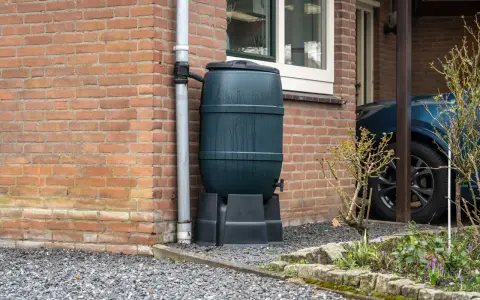 Zbiornik na wodę opadową przy ceglanym domu, zbierający deszczówkę z rynny. Ekologiczne rozwiązanie dla ogrodu.