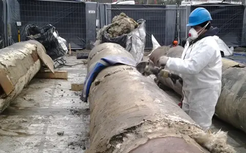 Pracownik w kombinezonie ochronnym usuwa izolację z rur, prawdopodobnie zawierającą eternit.