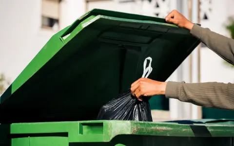 Ręka wrzuca worek ze śmieciami do zielonego kontenera. Zastanawiasz się, ile kosztuje wywóz śmieci?
