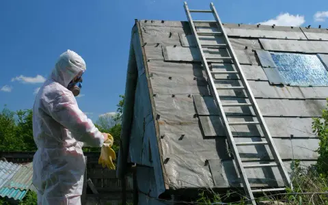 Osoba w kombinezonie ochronnym i masce przygotowuje się do prac związanych ze składowaniem eternitu na posesji. Na dachu widoczna drabina.