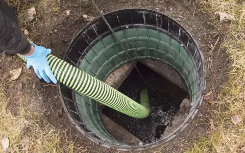 Ręka w niebieskiej rękawiczce wprowadza wąż do szamba betonowego. Drenaż szamba jest kluczowy dla prawidłowego funkcjonowania.