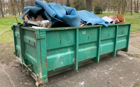 Zielony kontener KP7 wypełniony po brzegi niebieską wykładziną i innymi odpadami.