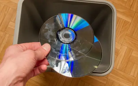 Dłoń trzyma pękniętą płytę CD/DVD nad szarym koszem na śmieci.