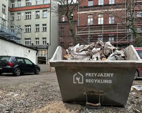 Kontener na odpady budowlane, kod odpadu 17 09 04. Wypełniony gruzem i opakowaniami, obok stoi czarny samochód.