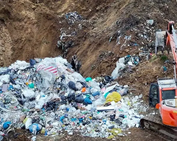 Ogromna góra śmieci, tworząca nielegalne wysypisko, z koparką w tle. Widać worki, folie i różne odpady.