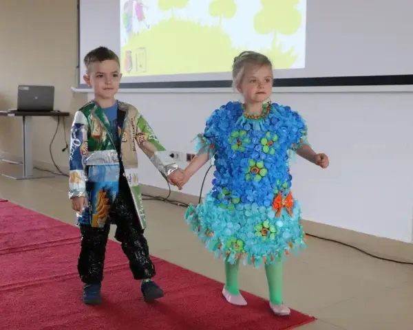 Chłopiec prezentuje strój z recyklingu, wykonany z opakowań po przekąskach. Jego koleżanka ma sukienkę z nakrętek.