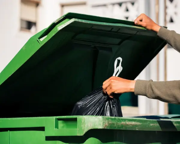 Ręka wrzuca worek ze śmieciami do zielonego kontenera. Zastanawiasz się, ile kosztuje wywóz śmieci?
