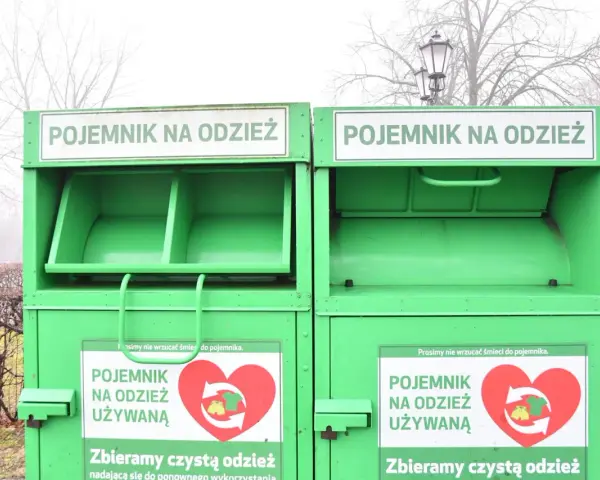 Dwa zielone kontenery na odzież używaną, z napisem "POJEMNIK NA ODZIEŻ UŻYWANĄ" i sercem z symbolami recyklingu.