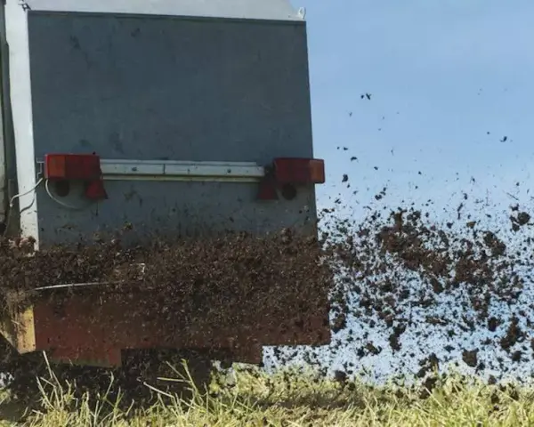 Rozrzutnik rozsypuje komunalne osady ściekowe na polu. Materiał jest wyrzucany w powietrze, tworząc brązową chmurę.