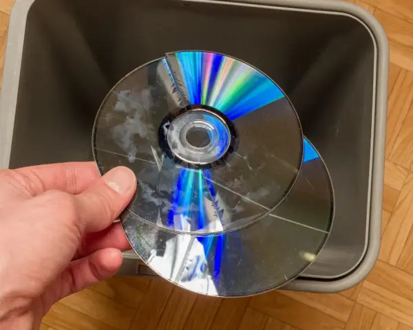 Dłoń trzyma pękniętą płytę CD/DVD nad szarym koszem na śmieci.