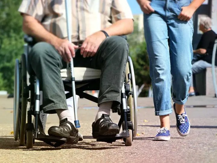 Osoba na wózku inwalidzkim z laską obok idącej osoby. Ulga na śmieci dla niepełnosprawnych to ważny temat.