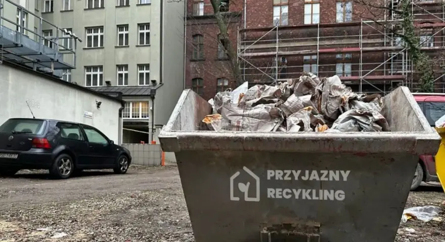 Kontener na odpady budowlane, kod odpadu 17 09 04. Wypełniony gruzem i opakowaniami, obok stoi czarny samochód.