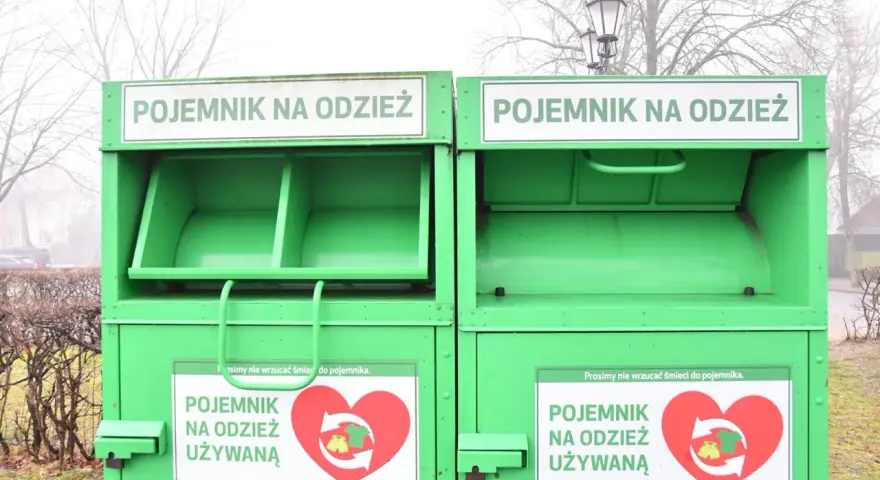Dwa zielone kontenery na odzież używaną, z napisem "POJEMNIK NA ODZIEŻ UŻYWANĄ" i sercem z symbolami recyklingu.