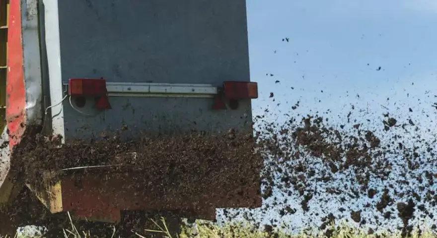 Rozrzutnik rozsypuje komunalne osady ściekowe na polu. Materiał jest wyrzucany w powietrze, tworząc brązową chmurę.