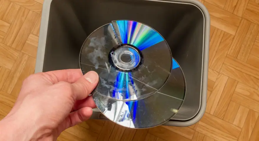 Dłoń trzyma pękniętą płytę CD/DVD nad szarym koszem na śmieci.