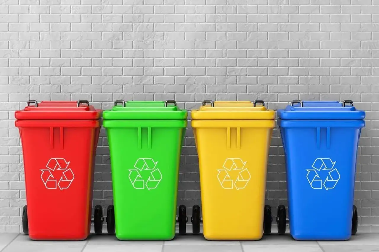 Cztery kolorowe kosze na śmieci (czerwony, zielony, żółty, niebieski) z symbolami recyklingu, gotowe do segregacji różnych rodzajów odpadów.