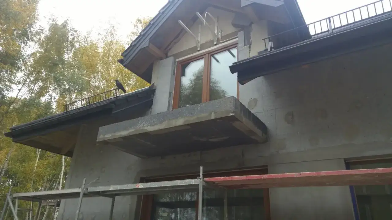 Budynek w trakcie prac wykończeniowych, widoczna termoizolacja, balkon i okna. Na dachu siedzi ptak.