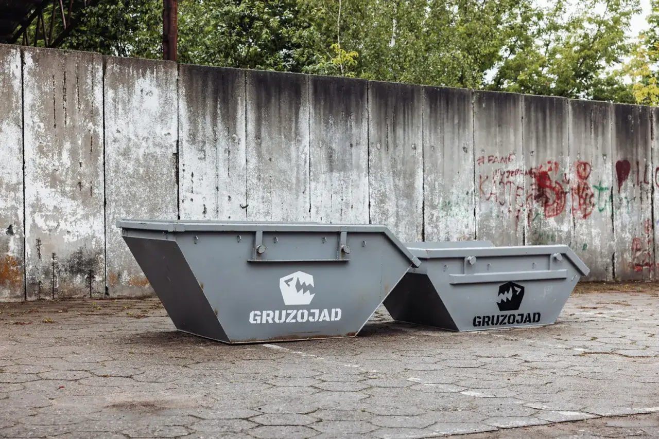 Dwa szare kontenery na gruz z logo GRUZOJAD stoją przed betonowym murem z graffiti. Numer 170107.