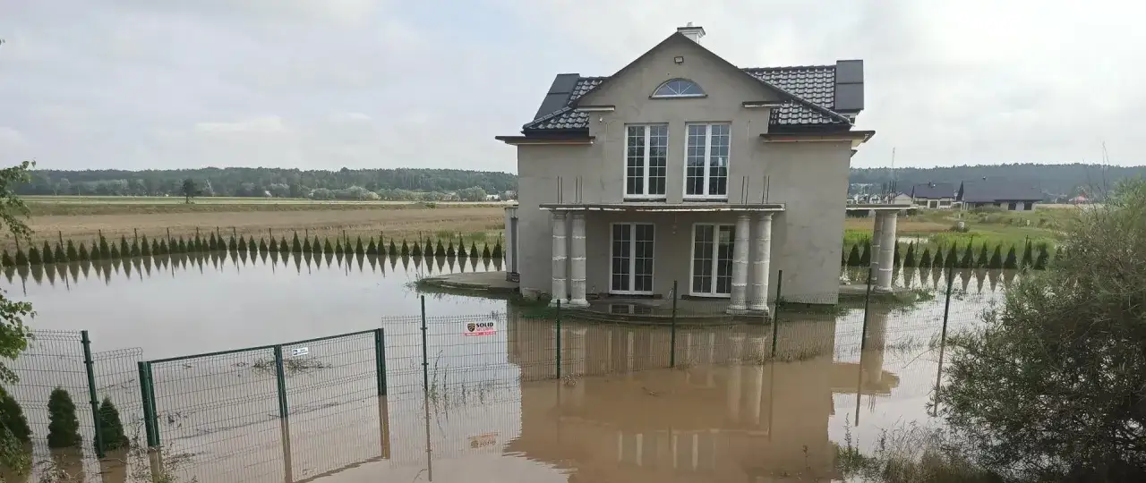 Dom otoczony wodą po powodzi błyskawicznej. Woda sięga do połowy ogrodzenia i zalewa teren wokół budynku.