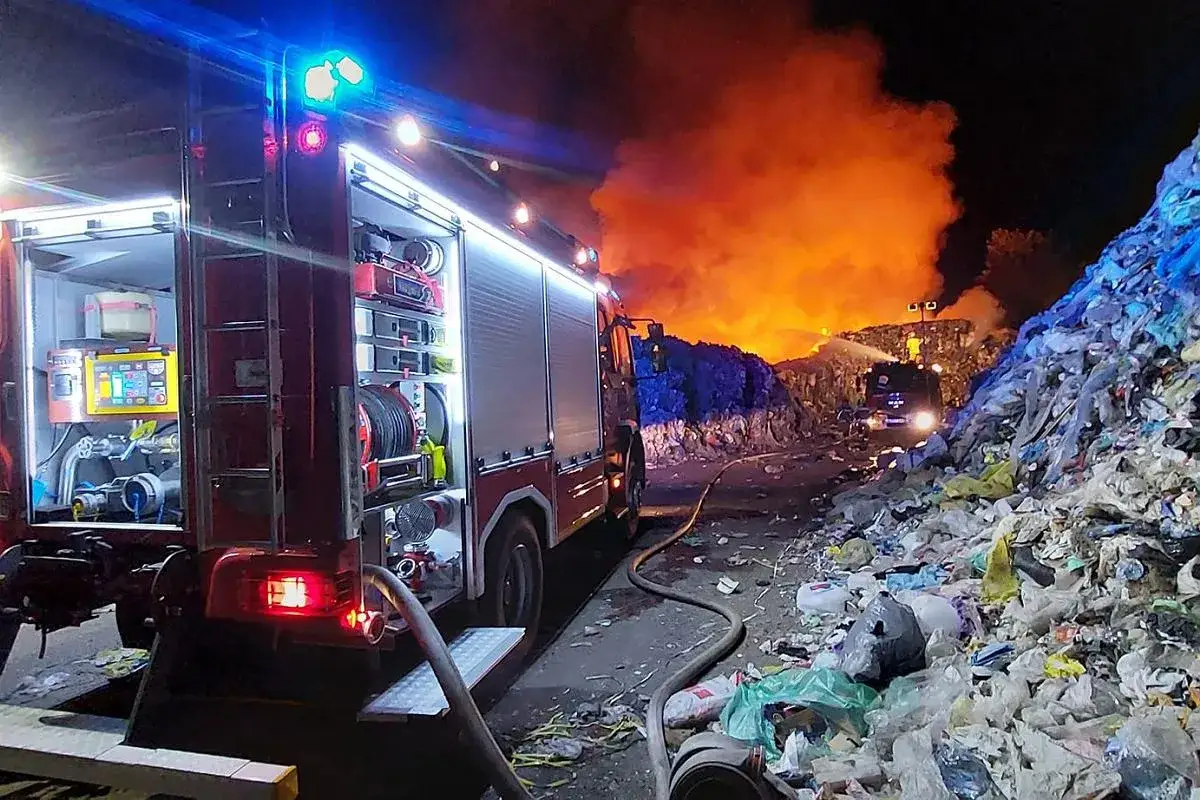 Straż pożarna walczy z pożarem wysypiska. Widać wóz strażacki i ogromny słup ognia.