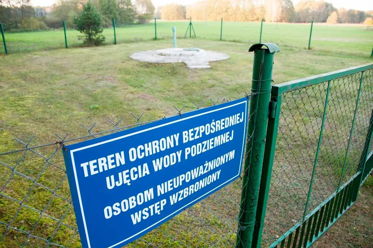 Niebieska tablica informuje o strefach ochronnych ujęcia wody podziemnej. Teren ogrodzony, wstęp wzbroniony.