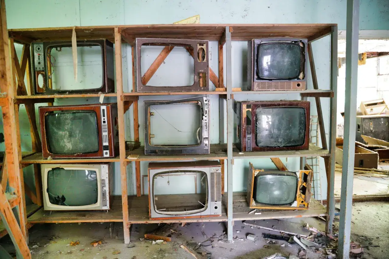 Stare telewizory na półkach, jak w miejscu, gdzie oddać stary telewizor. Wiele z nich jest uszkodzonych, z pustymi obudowami lub pękniętymi ekranami.