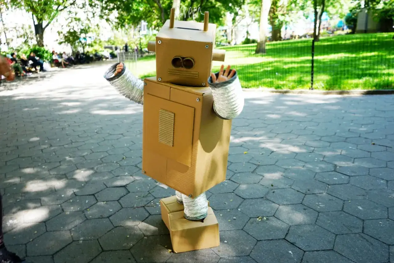 Chłopiec w stroju robota z recyklingu, z tekturowym korpusem i metalowymi rękawami, pozuje na tle parku.