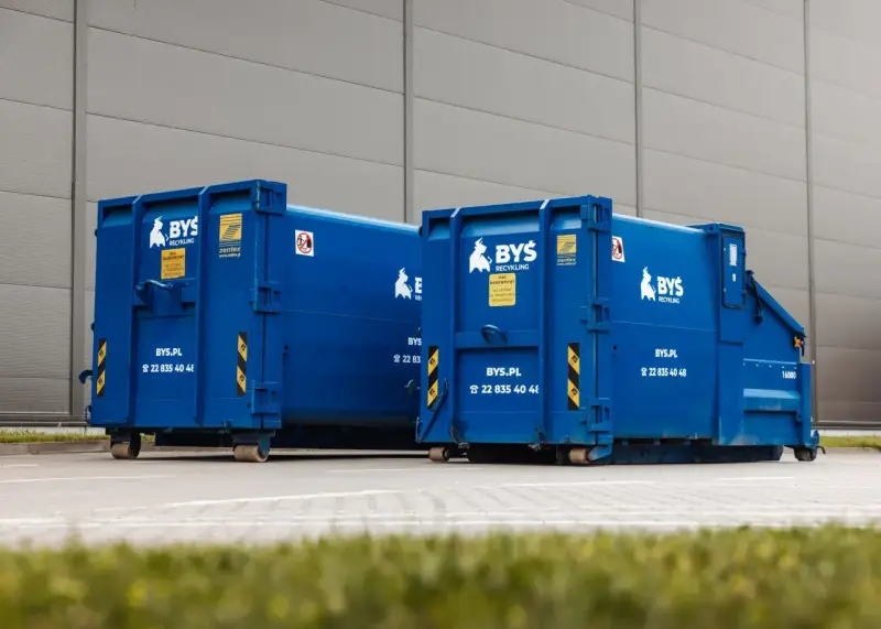 Dwa niebieskie kontenery BYS Recykling stoją przed budynkiem. Dowiedz się, ile kosztuje tona gruzu betonowego i jak go zamówić.
